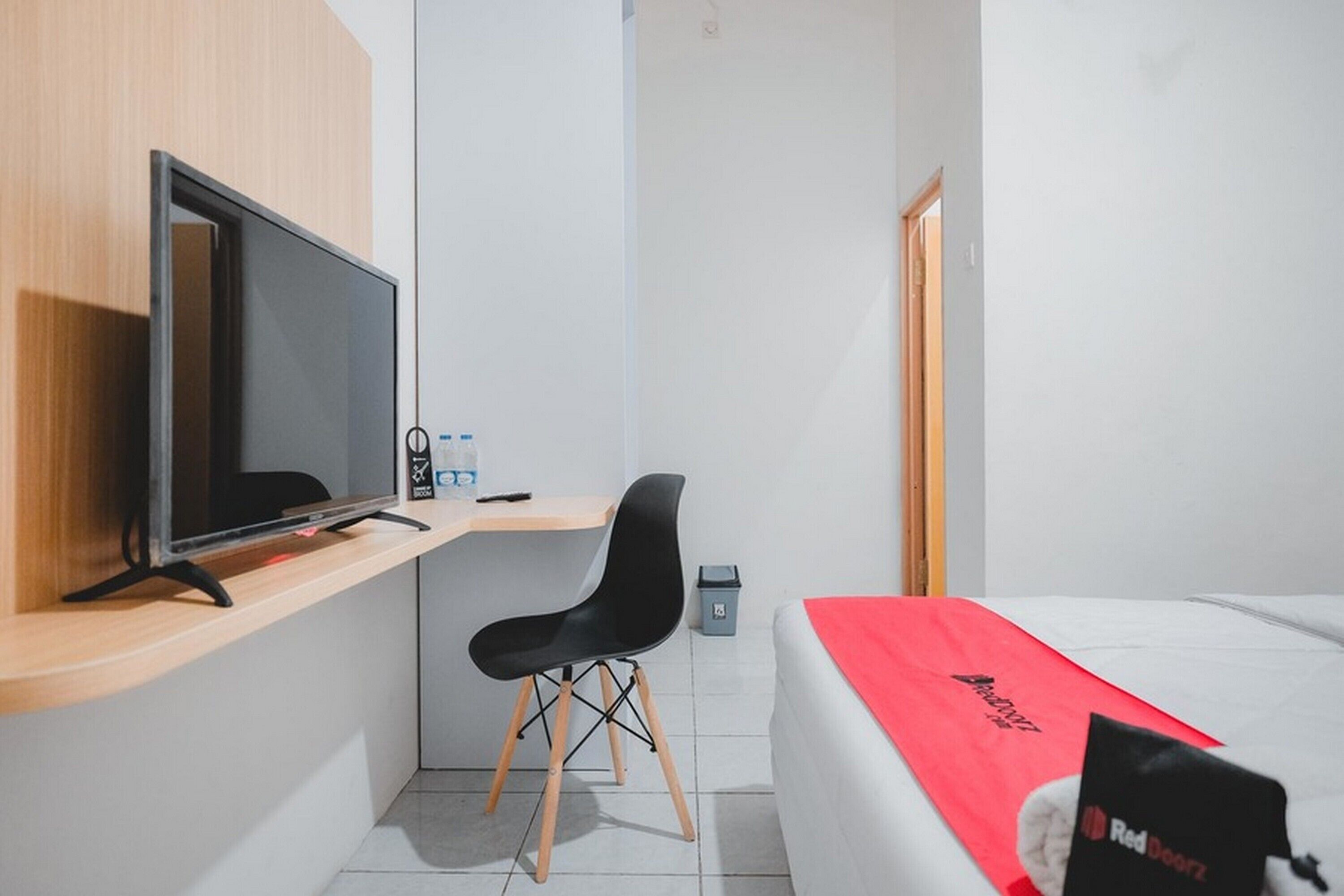 A small, modern hotel room with a bed, a desk with a computer monitor, a chair, and a red bedspread.