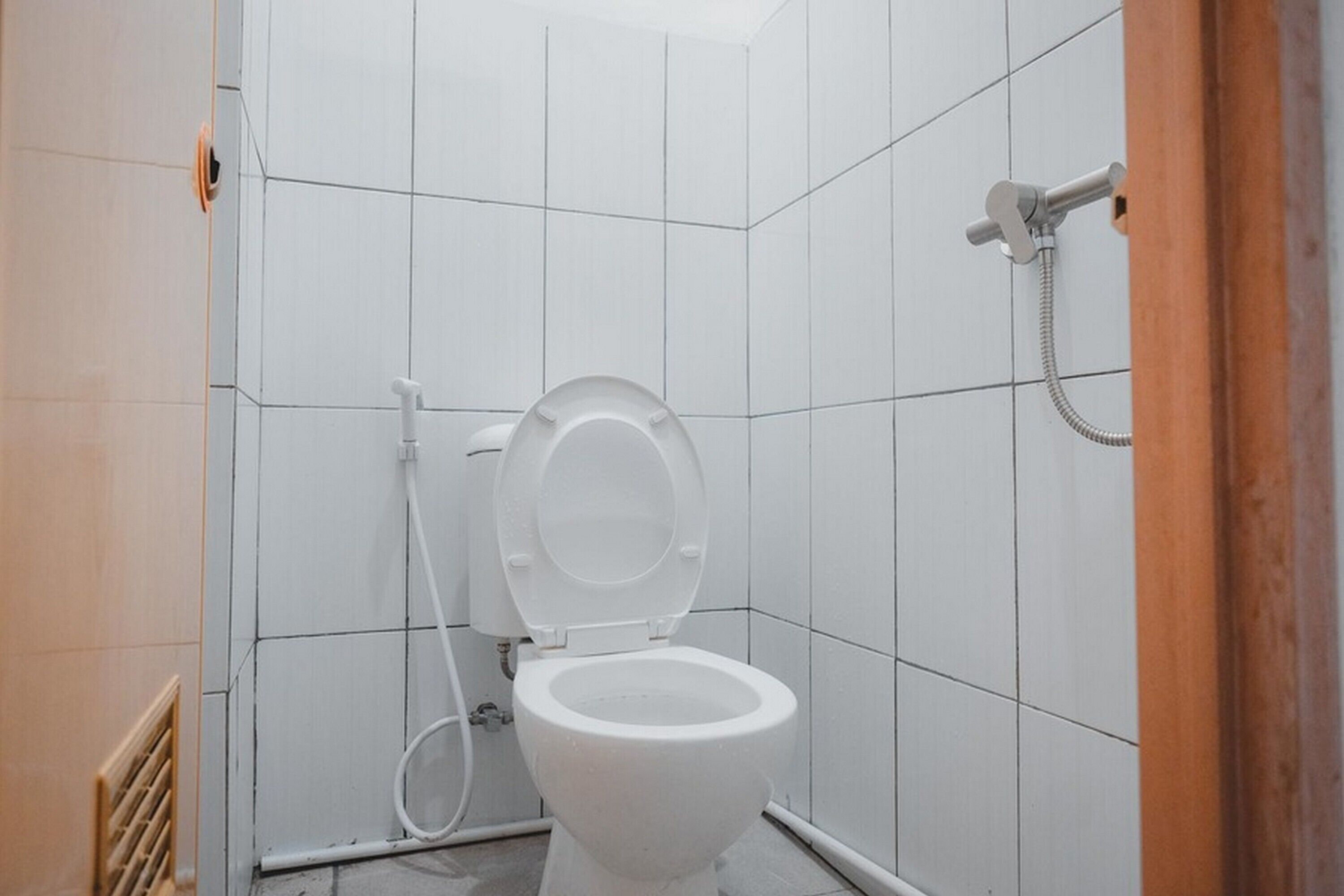 A small, white-tiled bathroom with a toilet, a shower, and a visible vent on the left.