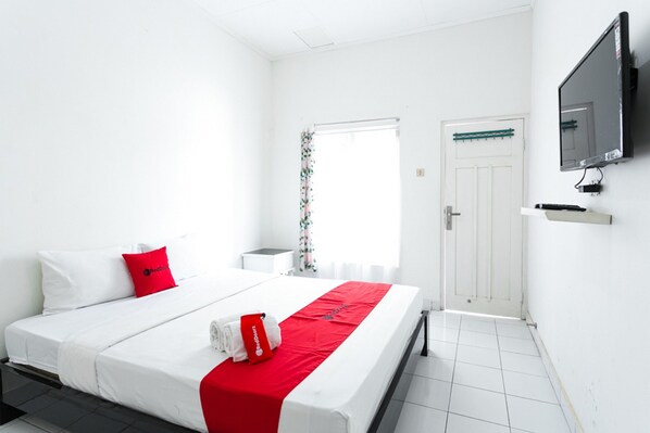 Quarto de hotel com uma cama, televisão fixada na parede, janela com cortinas e uma porta.