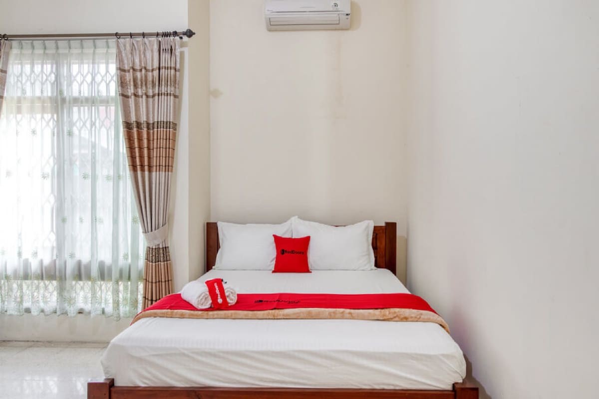 A neatly made bed with white bedding and a red pillow, a wooden headboard, and a window with sheer curtains.