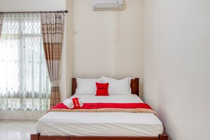 Una cama bien hecha con ropa de cama blanca, un almohadón y una colcha rojos. A la izquierda, hay una ventana con cortinas de encaje y un cortinaje estampado. Una unidad de aire acondicionado está montada en la pared sobre la cama.
