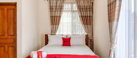 Quarto de hotel com cama, manta vermelha, travesseiro e janela com cortinas.