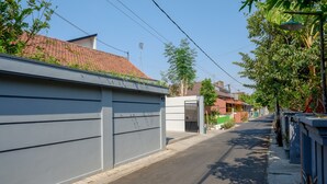 Eine ruhige Wohnstraße mit einer grauen Wand auf der linken Seite, einem kleinen Verteilerkasten und einer Reihe Häuser mit roten Ziegeldächern.