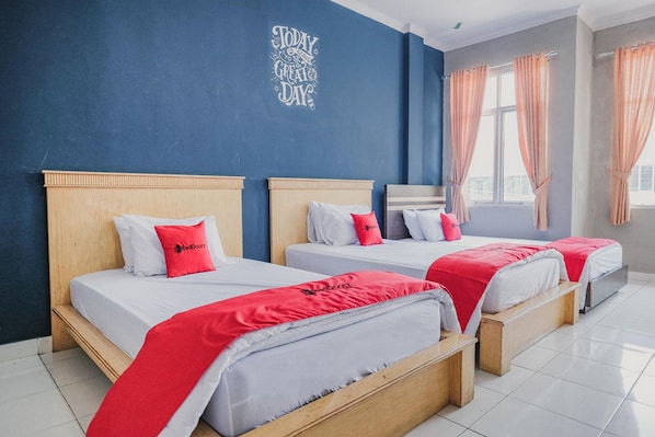 A hotel room with three beds, each with a wooden headboard and white bedding. There is a blue accent wall with a positive message, and the room has large windows with curtains.