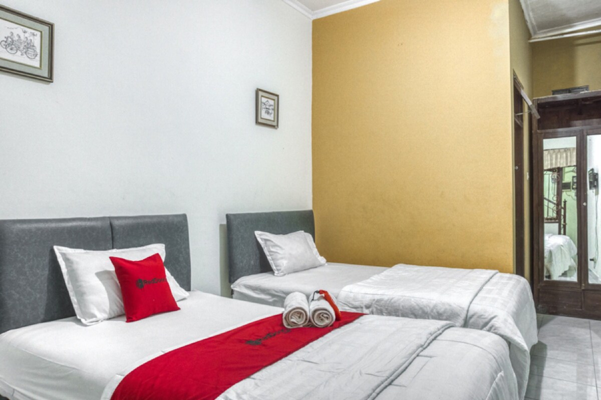 A hotel room with two beds, white bedding, red and white pillows, a red throw blanket, and a pair of rolled towels.