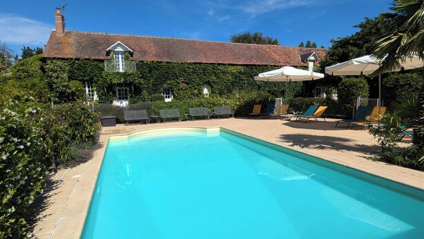 A swimming pool with lounge chairs and umbrellas, surrounded by lush greenery and a brick building with a tiled roof.
