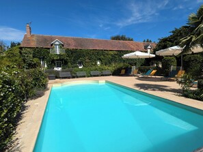 Une piscine entourée de chaises longues et de parasols, elle-même bordée d’une végétation luxuriante et d’un bâtiment en briques surmonté d’un toit en tuiles.