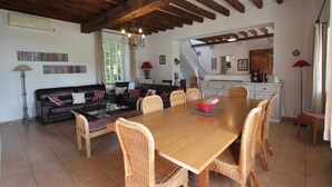 A spacious living area with a wooden dining table and wicker chairs, a dark leather sofa, and a staircase leading to an upper level.
