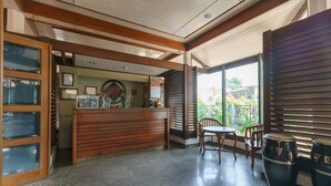 A reception area with a wooden counter, a small table with chairs, and a view of the outdoors through large windows. - RedDoorz near Transmart Pangkal Pinang (Pangkalpinang)