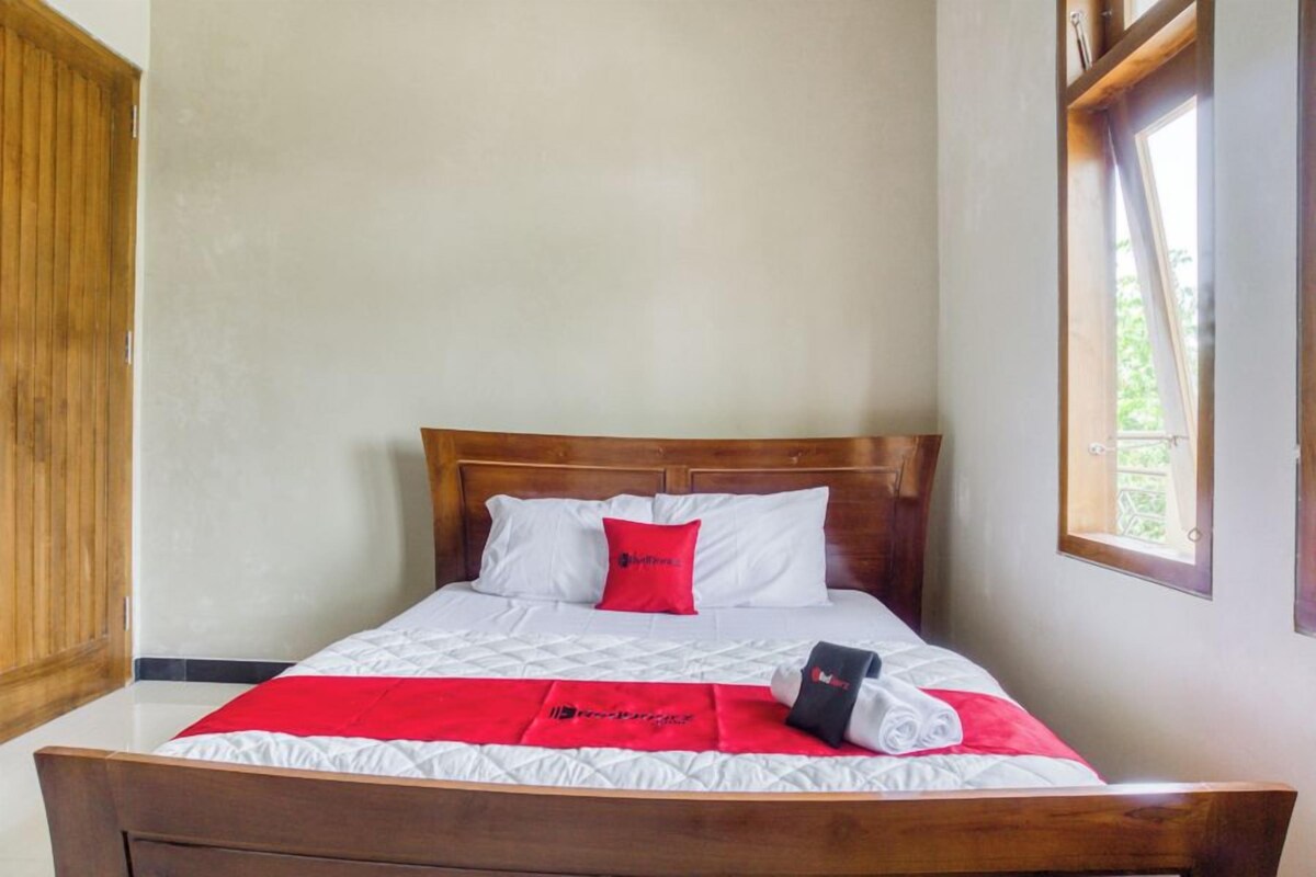 Un lit bien fait, avec une tête de lit en bois, une literie blanche et un coussin rouge qui apporte une touche de couleur.