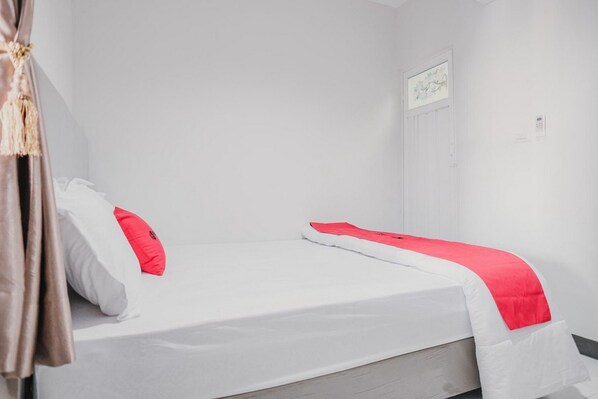 Una cama individual con ropa de cama blanca y una almohada roja decorativa, un cabecero de madera y una pared blanca con un pequeño cuadro enmarcado.