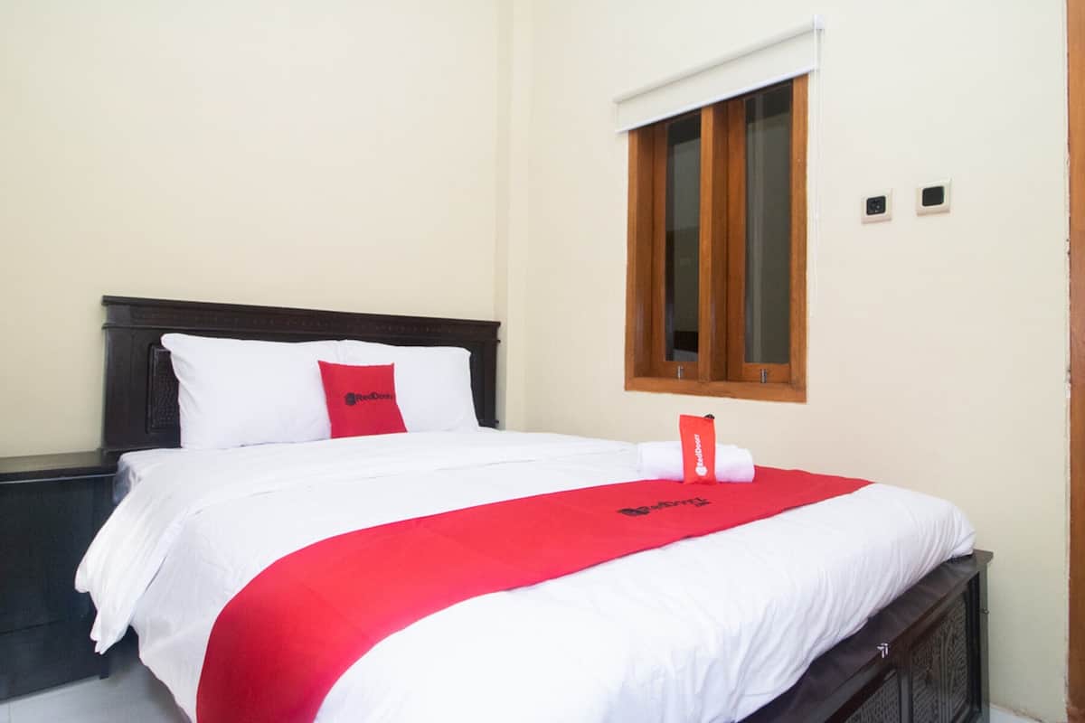 A neatly made bed with white and red bedding, a wooden headboard, and a window with wooden blinds.