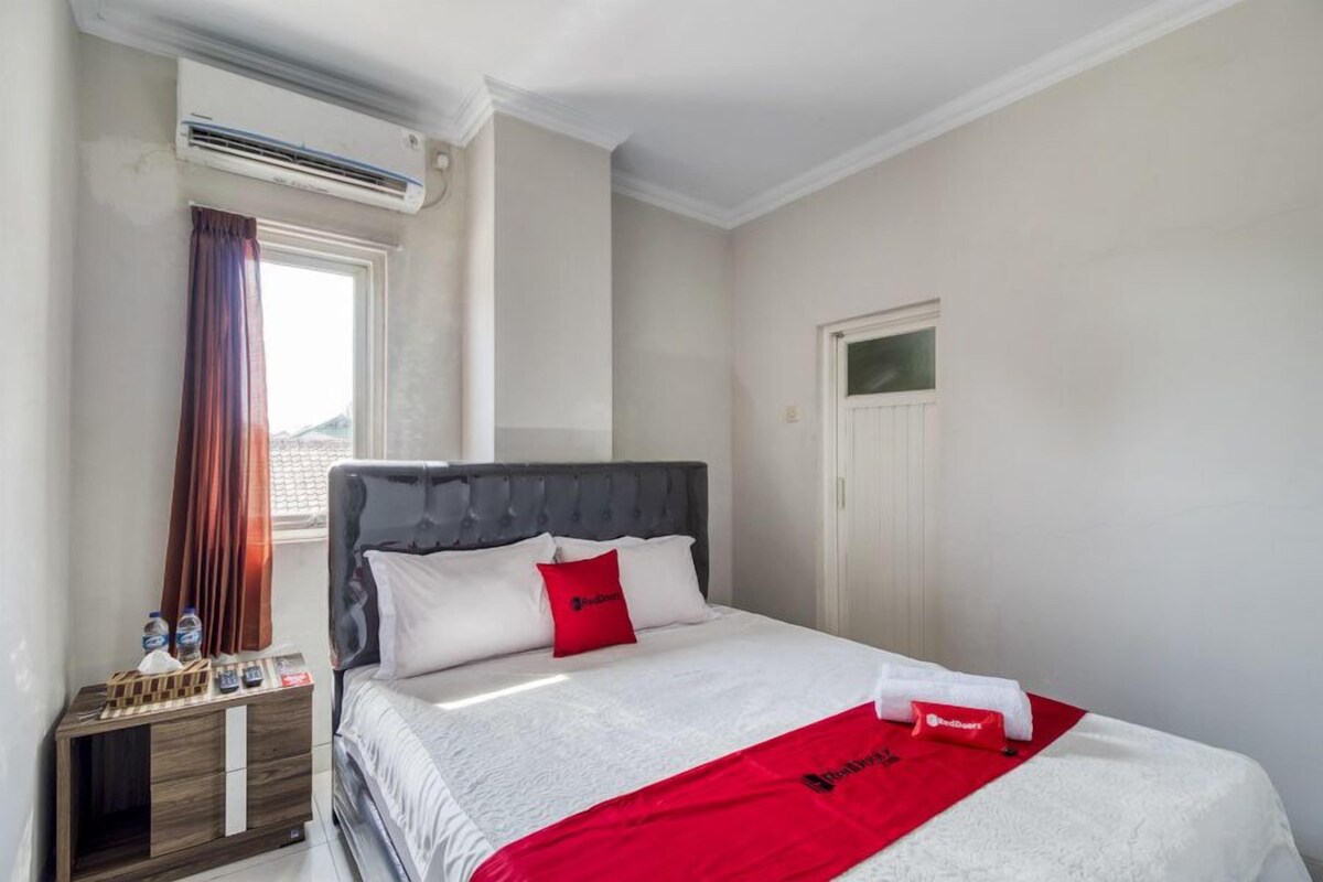 A neatly arranged hotel room with a bed, a bedside table with a lamp, a window with curtains, and an air conditioning unit.
