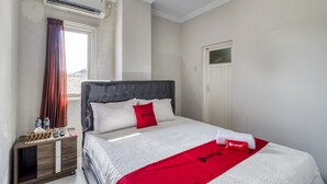 A hotel room with a bed, a nightstand, an air conditioning unit, a window with curtains, and a door. - RedDoorz near Condongcatur Bus Station (Depok)
