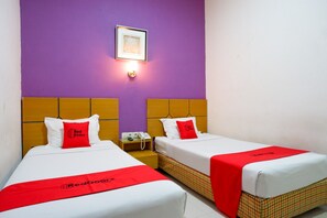 A hotel room with two beds, each with a red bedspread and pillow, a small bedside table with a phone, and a wall-mounted light fixture.