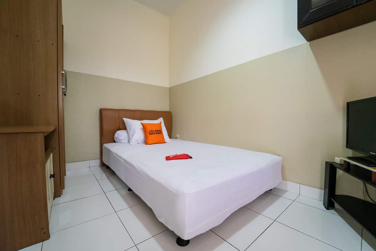 Una cama bien hecha con ropa blanca y un almohábil naranja, un armario de madera y un televisor sobre un mueble.