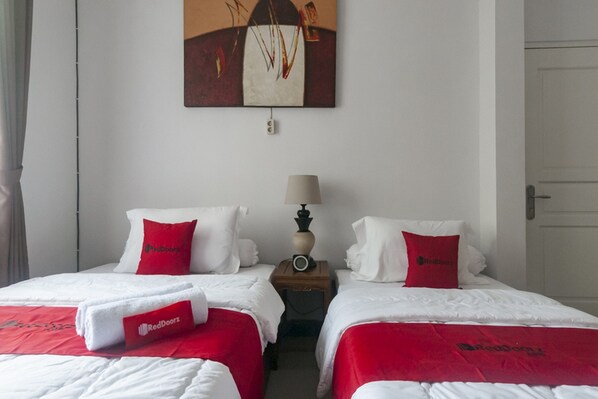 Habitación de hotel con dos camas, cada una con ropa blanca y cabeceros rojos. Hay un cuadro en la pared sobre las camas, una mesita de noche con una lámpara y una puerta a la derecha.