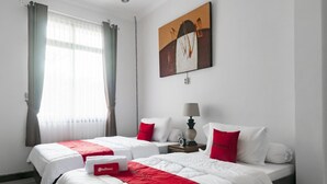 Une chambre d’hôtel avec deux lits, une literie rouge et blanche, un tableau au mur et une fenêtre avec des rideaux.
