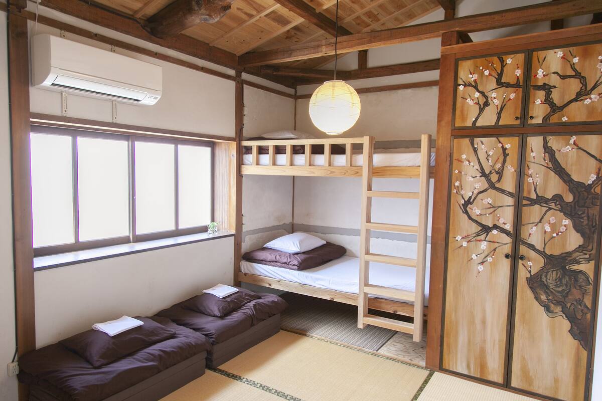 Une pièce avec un lit superposé, un futon, un revêtement de sol en tatami, une porte coulissante ornée de motifs de fleurs de cerisier et un ventilateur de plafond.