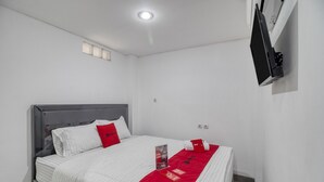 A neatly made bed with white and red bedding, a flat-screen TV mounted on the wall, and a small window with blinds. - RedDoorz Syariah near Galeria Mall Yogyakarta (Yogyakarta)