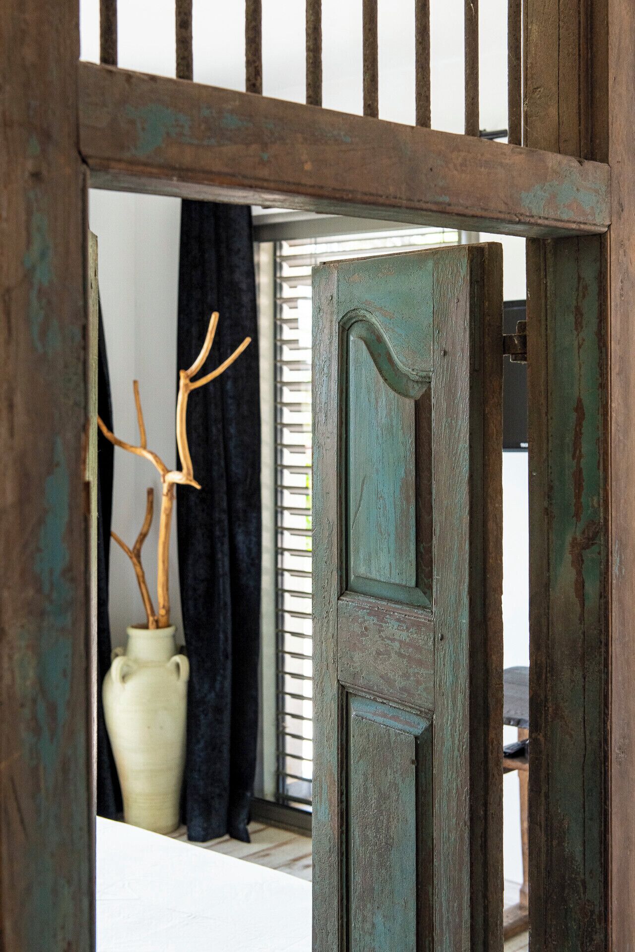 A rustic wooden door with peeling paint, a white vase with branches, and a dark curtain.