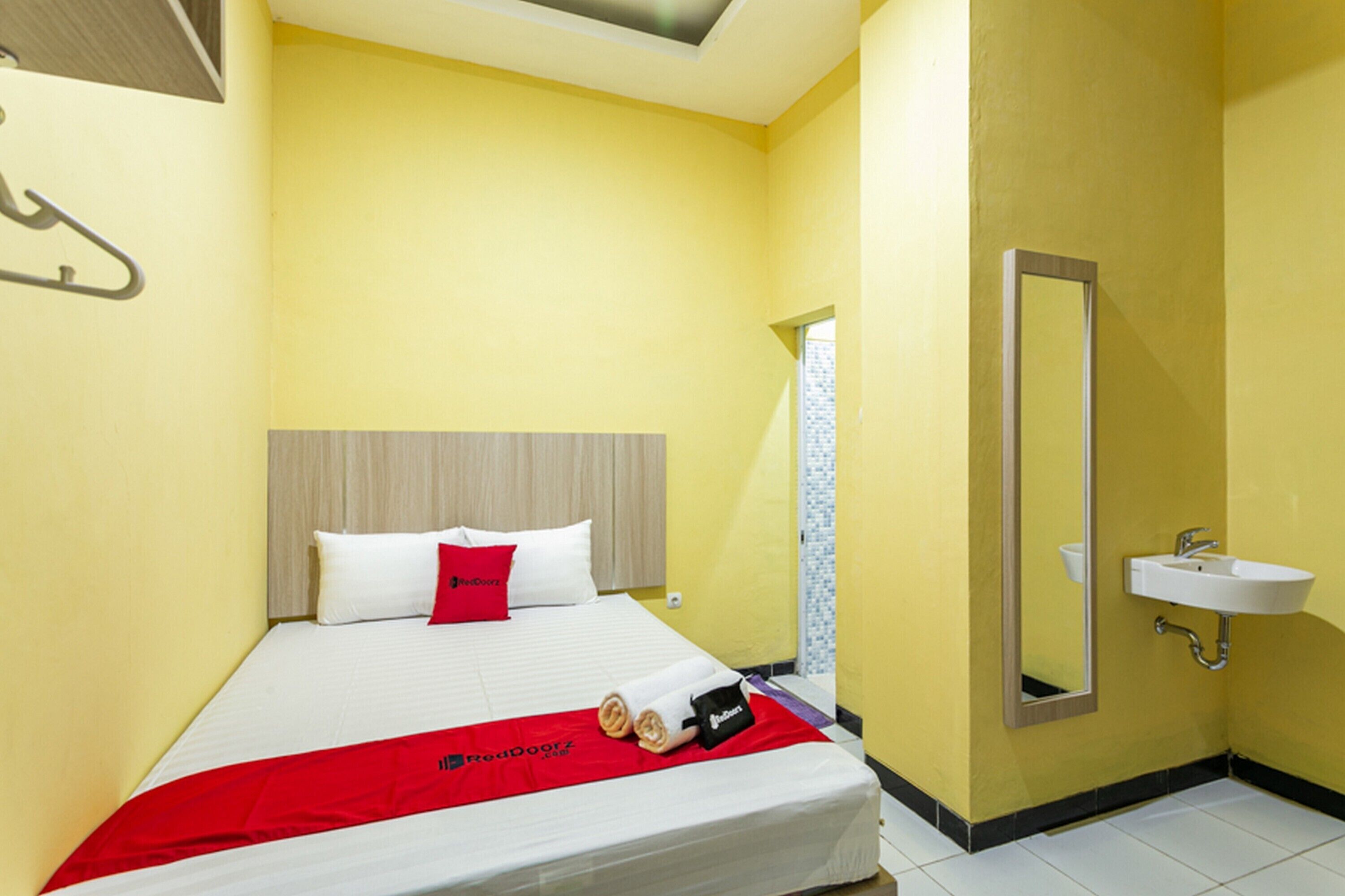 A hotel room with a bed, a red blanket, a white pillow, a small black bag, and a bathroom with a sink and mirror.
