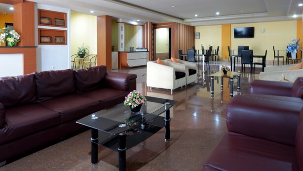 Een hotel lobby met een bruine leren bank, een witte bank, een glazen salontafel met een bloevas, en een eetgedeelte met stoelen en tafels.