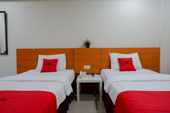Une chambre d’hôtel avec deux lits, chacun agrémenté d’un oreiller rouge et de draps blancs. Une petite table avec un téléphone et un vase de fleurs se trouve entre les deux lits.