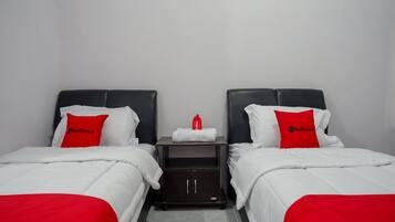 Habitación de hotel con dos camas, cada una con ropa blanca y almohadas rojas. Hay una pequeña mesa con una toalla blanca y un objeto rojo entre las camas.