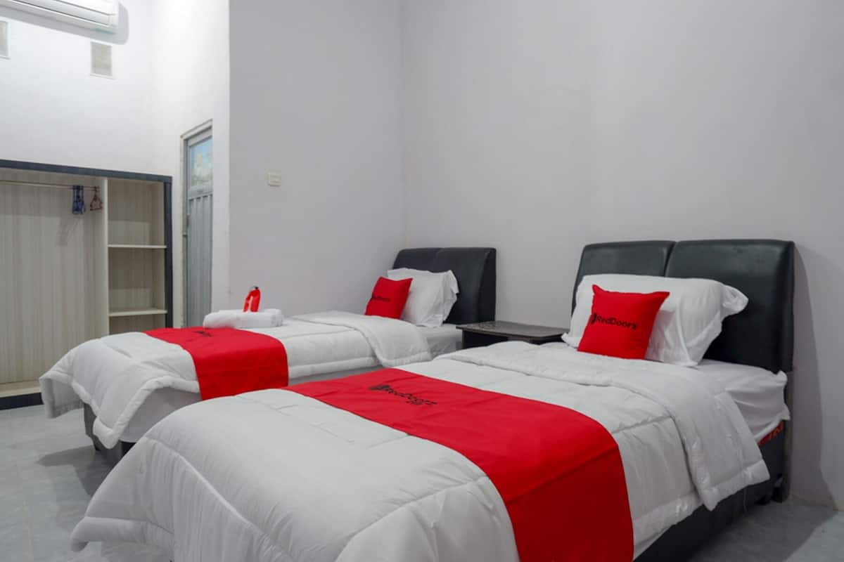 A hotel room with two beds, each with white bedding and red accents, a wardrobe, and a small table.