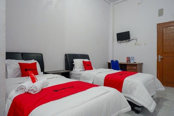 Une chambre d’hôtel avec deux lits, chacun agrémenté de draps blancs et de touches de couleur rouge, un petit bureau avec une chaise, un téléviseur fixé au mur et une porte en bois.