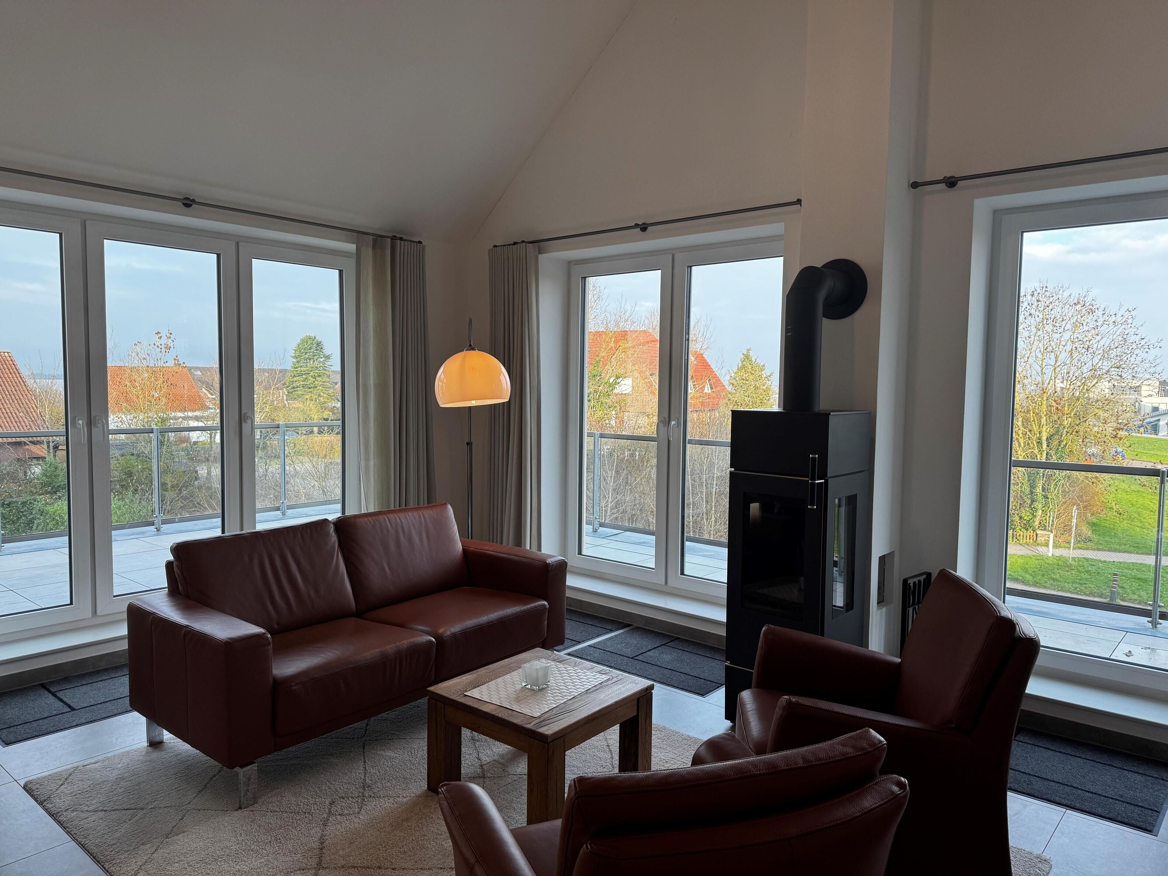 Ein geräumiges Wohnzimmer mit einer braunen Ledersofa, einem Holztisch und einem schwarzen Kamin. Große Fenster bieten Blick auf die Nachbarschaft.