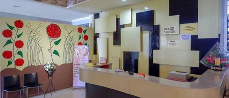 A hotel reception area with a reception desk, two chairs, a wall with floral artwork, and a fire extinguisher.