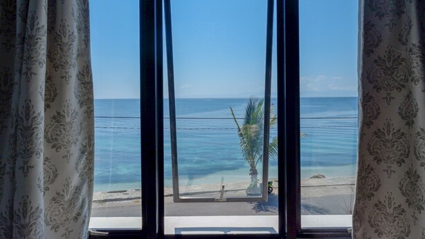 A view through a window with curtains, featuring a beach, palm tree, and clear sky. - RedDoorz Plus Syariah @ Hotel Boulevard Luwuk (Luwuk)