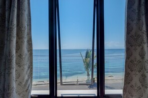 Une vue à travers une fenêtre avec des rideaux, donnant sur une plage, un palmier et un ciel dégagé.