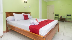 A neatly made bed with a red and white bedspread, a wooden headboard, and a red pillow. There is a small bedside table with a lamp, a chair, and a door in the background. - RedDoorz @ Frans Seda Kupang (Kupang)