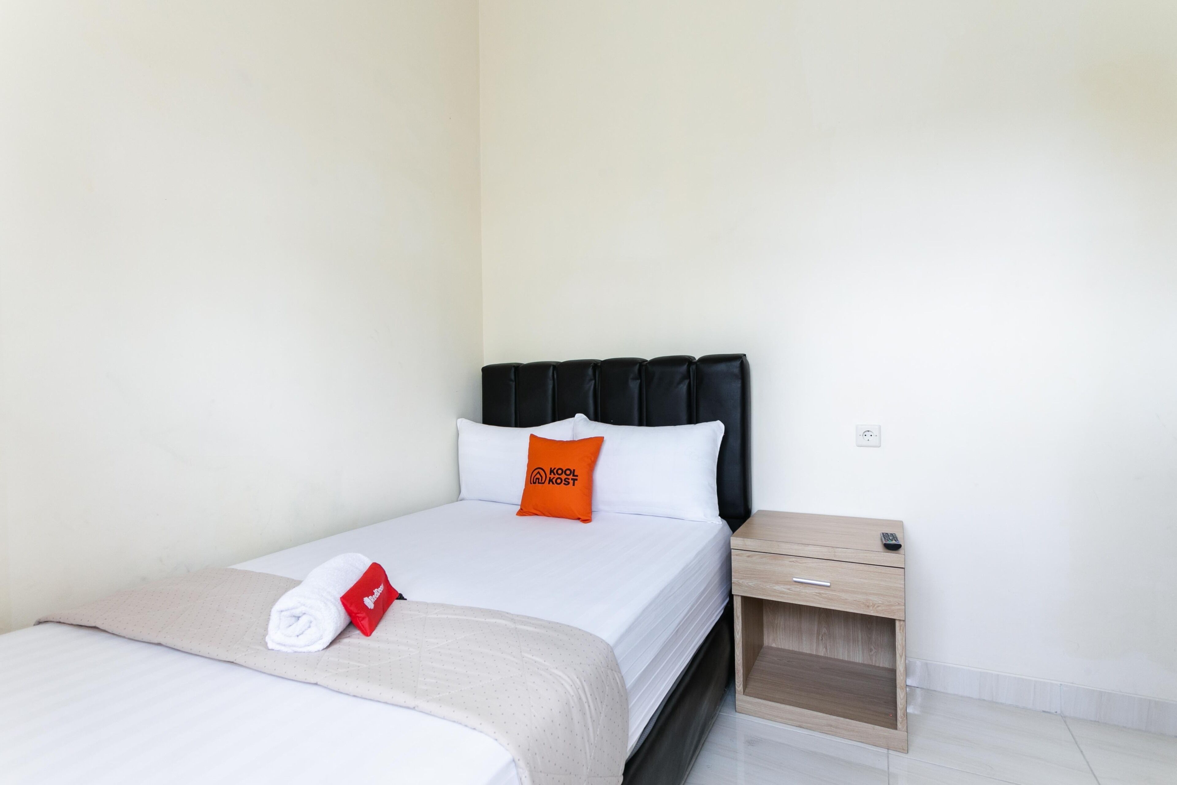 A neatly made bed with a white comforter and a red pillow, a wooden nightstand with a remote control, and a light-colored wall.