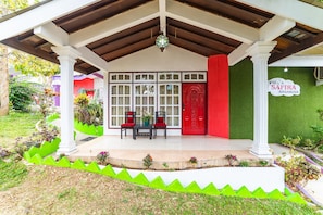 A covered outdoor area with a red door, a white sign reading "Villa Safira Basanova", and a seating arrangement with two chairs and a table.