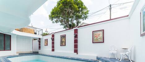 A swimming pool with blue and white tiles, a tree, and a white wall with framed pictures.