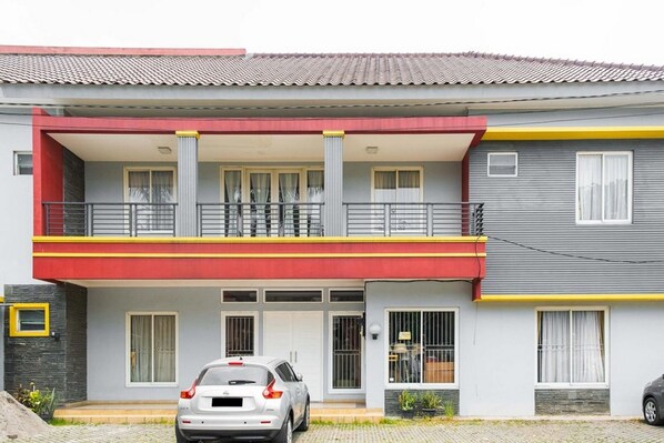 A two-story residential building with a balcony, a car parked in front, and a small garden area.