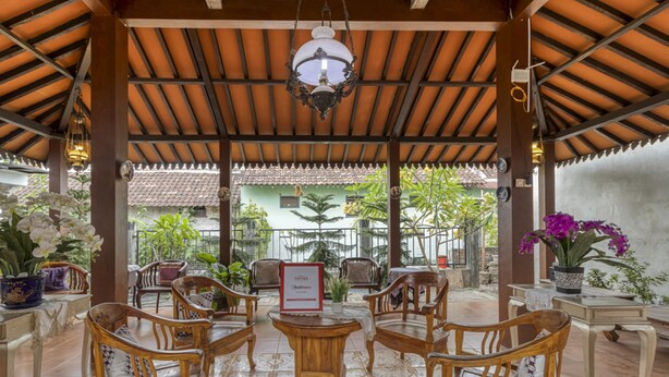 A covered outdoor seating area with wooden chairs and tables, surrounded by potted plants and a view of a building with a tiled roof. - RedDoorz Syariah Near Wijilan 2 Yogyakarta (Yogyakarta)