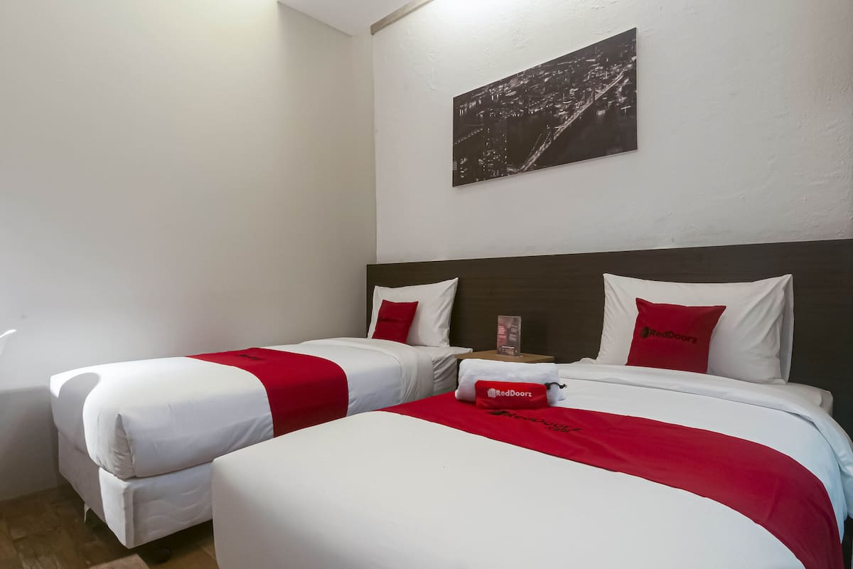 Camera d'albergo con due letti, entrambi con biancheria bianca e dettagli rossi, un comodino con una lampada e un dipinto raffigurante una vista cittadina sulla parete.