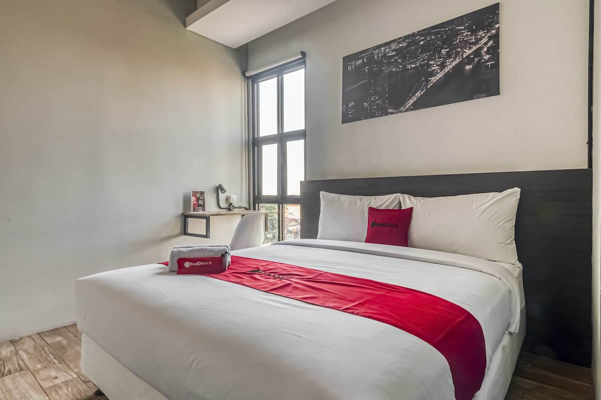 Una camera d'hotel con un letto grande, una coperta rossa, un cuscino rosso, un comodino con una lampada e un dipinto che raffigura un panorama urbano.