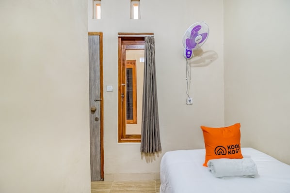 A hotel room with a bed, a pillow with "Kool Kost" logo, a wall fan, and a window with curtains.
