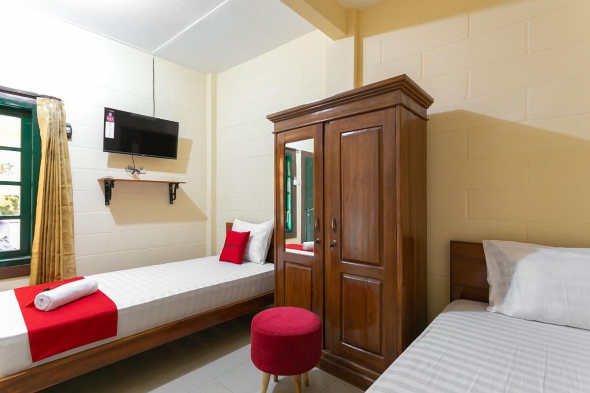 A room with two single beds, a wooden wardrobe, a television mounted on the wall, and a window with curtains.