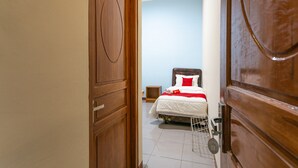 Habitación de hotel con puerta de madera, cama con ropa blanca y detalles rojos, y una mesita de noche pequeña.