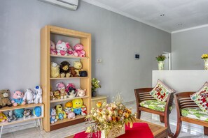 Une pièce avec une bibliothèque en bois remplie de peluches, une chaise en bois avec des coussins à motifs et un vase de fleurs posé sur une table rouge.