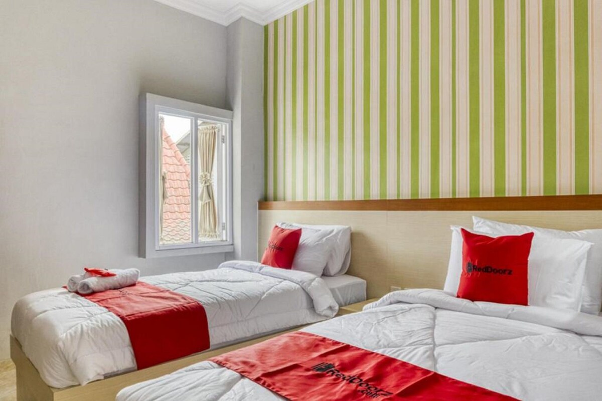 A hotel room with two beds, each with white bedding and red accents, a striped green and white wall, and a window with a view of buildings.