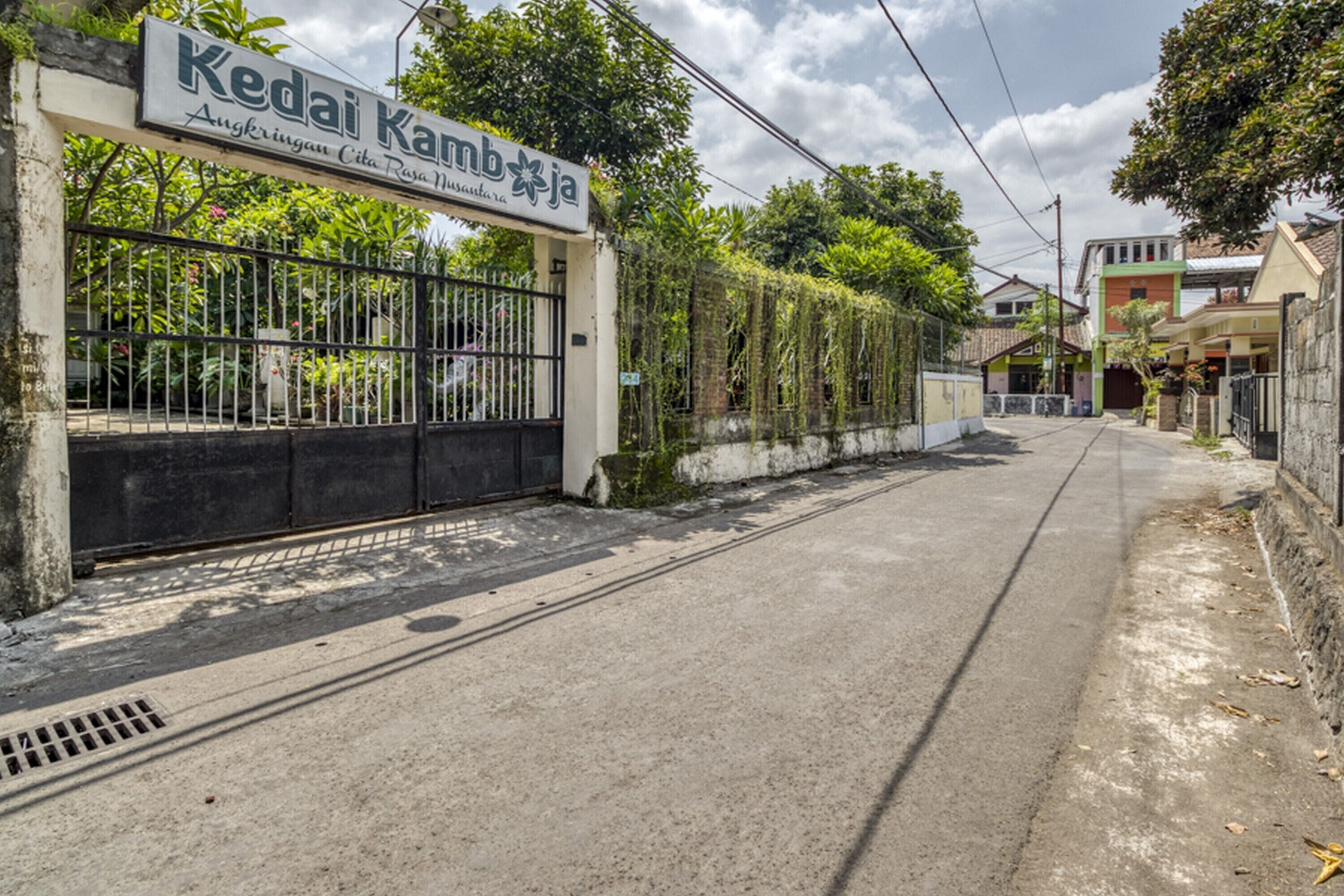 A gated entrance with a sign reading "Kedai Kambang" and a pathway leading into a residential area.