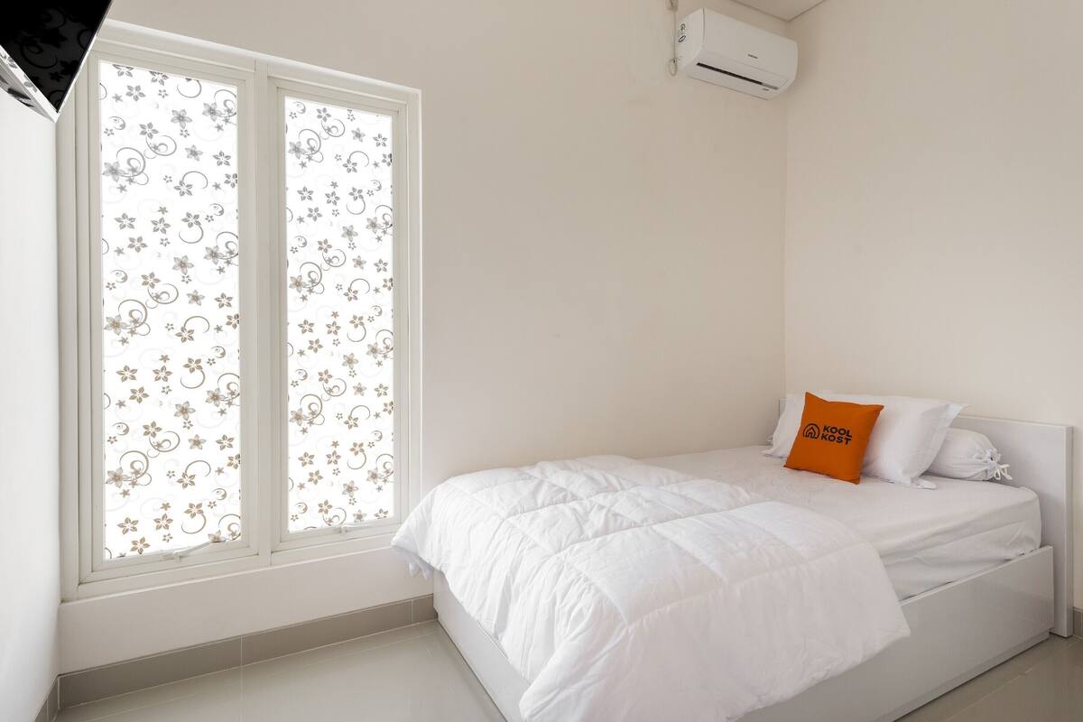 A bedroom with a bed, a window with floral blinds, and an air conditioning unit.